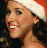 A woman wearing a red and white Santa hat smiles with her mouth open, showing teeth. The background is dark and textured.