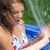 A woman in a colorful bikini stands near water, with green grass and blue pool visible behind her