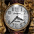 A circular clock face with black numerals and hands on a light gray background, set against a brown textured backdrop.
