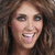 A static pixelated image of a smiling person with long brown hair, visible teeth, and dark eye makeup against a blurred background.