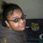 A person with dark hair in a bun wears sunglasses and a gray hoodie, positioned against a blurred background with indistinct shapes.