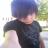Pixelated static image of a person with short dark hair wearing a dark shirt, standing outdoors with blurred greenery and light background.