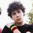 Pixelated image of a person with dark curly hair wearing a black top with colorful stripes on the sleeve, next to a blue background with pixelated text.