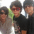 Three individuals in dark clothing stand closely together, one wearing red-tinted glasses and a green accent on the left side of the image.