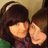 Two people with dark hair and bangs pose closely together, one in a green top and the other in purple, against a light brown background.