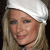Pixelated close-up of a person with light brown hair wearing a white head covering and dark clothing against a dark background.