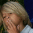 A blonde woman in a white top covers her mouth with her hand, her eyes closed, against a dark blue background with green foliage.