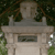 A stone monument with a rectangular base and vertical pillars, set against a backdrop of green foliage and brown branches.