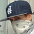 Pixelated close-up of a person wearing dark blue cap with white 'L' logo, brown hair visible beneath brim, light-colored shirt, and sunglasses on face.