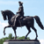 Pixelated image of a dark horse statue with a rider holding a raised sword, standing on a green base against a light blue sky.