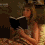 A woman in a sleeveless dress holds a black book with white pages, then lowers it to reveal a small dark object in her hand.