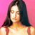 Pixelated image of a person with long dark hair against a solid pink background, wearing a light-colored top with red straps.