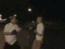 Two figures in light shirts and dark pants interact on a dimly lit street at night, with one figure raising arms while the other bends forward.