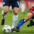 Pixelated static image of soccer players' legs and a ball on grass, with visible uniform colors and partial player poses.
