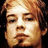Pixelated close-up of a person's face with closed eyes, light brown hair, and a neutral expression against a dark background.