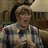 A person in a plaid shirt holds an object with both hands, seated near a lamp and bookshelf background.