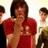 Red background with pixelated text and a central figure in red shirt making hand gesture, flanked by two blurred figures.