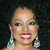 A pixelated close-up of a person with light skin, dark eyes, and red lips smiling, wearing large silver earrings against a dark background.