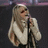Pixelated image of a person with long blonde hair holding a microphone, surrounded by red pixelated text on a bright pink background.