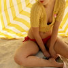 A person in a yellow striped crop top and red bikini bottoms sits on a sandy surface under a yellow and white striped canopy.