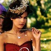 A woman in a red strapless dress with a white circular emblem, wearing a jeweled headpiece and heart-shaped necklace, holding a small object near her shoulder.