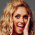 A person with long curly blonde hair wears a red shirt with white text and heart symbols, smiling with one hand near their face against a gray background.