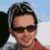 Pixelated static image of a person with dark hair wearing sunglasses and an orange shirt against a blue sky background.
