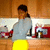 A pixelated static image shows a person with dark hair in a ponytail wearing a light gray top and bright yellow bottom, standing in front of a kitchen counter with red appliances.