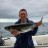 A pixelated image shows a person holding a large fish horizontally with both hands against a blurred beach background.