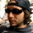 A person with light brown hair wears a dark beanie and dark sunglasses, facing left in a static image with a blurred background.