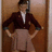 A pixelated static image shows a person standing with hands on hips against a brown door, wearing a dark jacket over a white shirt and light-colored pants.