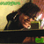 A person in a dark cap and jacket holds a green device, with a blurred background showing green text and a black bar.