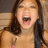 A pixelated close-up of a person with long dark hair, mouth open in an expressive shout or laugh, wearing a light-colored top against a blurred background.