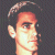 Pixelated close-up of a person's face with light skin tone, dark hair styled in a side part, and a neutral expression against a dark checkered background.