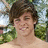 Pixelated image of a person with short brown hair smiling, set against green foliage and light sky background.