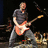 Pixelated image of a person playing an electric guitar, wearing a dark shirt and light pants against a dark background.