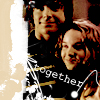 Two people in a close-up pose with a black background, one wearing a black shirt with 'together' text and a necklace, the other with light brown hair, both smiling.