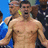 A pixelated image of a shirtless man with short dark hair and facial stubble, standing against a blue background with blurred figures behind him.