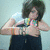 A person with short dark hair wears a dark sleeveless top and colorful beaded necklace, making a hand gesture with both hands near the chest against a light blue background.