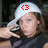 A pixelated image shows a person wearing a white cap with a red circular symbol containing a '3' on the front, holding a dark object near their face against a dark background.