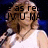 A woman with shoulder-length brown hair in a white sleeveless top holds a microphone against a dark blue background, with partial text overlays visible.