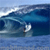 Pixelated static image of a surfer riding a large blue ocean wave with white foam, set against a light blue sky background.