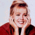 A pixelated image of a person with light brown hair in an updo, wearing a red jacket over dark clothing, resting hands on cheeks against a white background.