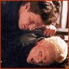 Two people embrace in a dimly lit indoor setting with one person resting their head on the other's shoulder.