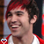 A person with red and dark brown hair wearing a white shirt under a red jacket, smiling broadly in with blue heart icons below