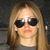 A pixelated close-up of a person with long light brown hair wearing dark sunglasses and a gray top against a dark background.