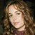 Pixelated close-up of a person with light brown curly hair, wearing a red garment, against a dark background.
