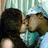 Two figures in a close embrace, one wearing a light blue cap and sleeveless top, the other with dark hair; background features vertical striped curtains.