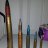 Pixelated vertical arrangement of six slender objects resembling pens or brushes against a muted gradient background.
