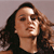 A pixelated close-up of a person with long dark hair, wearing a dark top, against a plain light brown background.